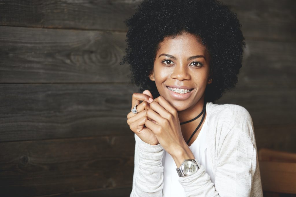 Curly-haired woman wearing braces and a white shirt smiles against a wooden wall