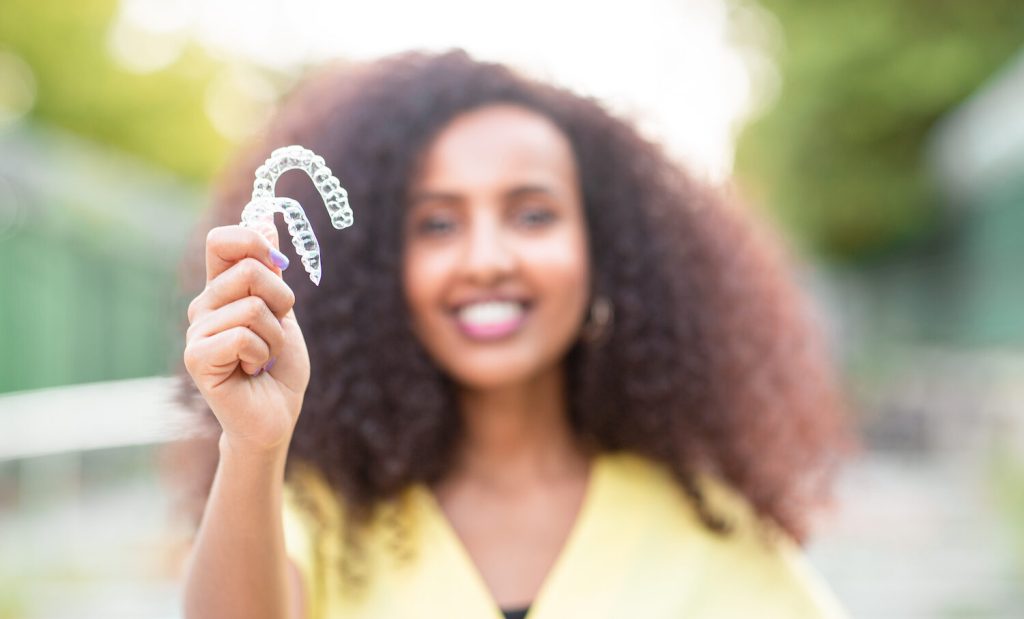 Blurred Black woman with big curly hair in a yellow blouse smiles while holding 2 Invisalign aligners outside on a walkway by some trees