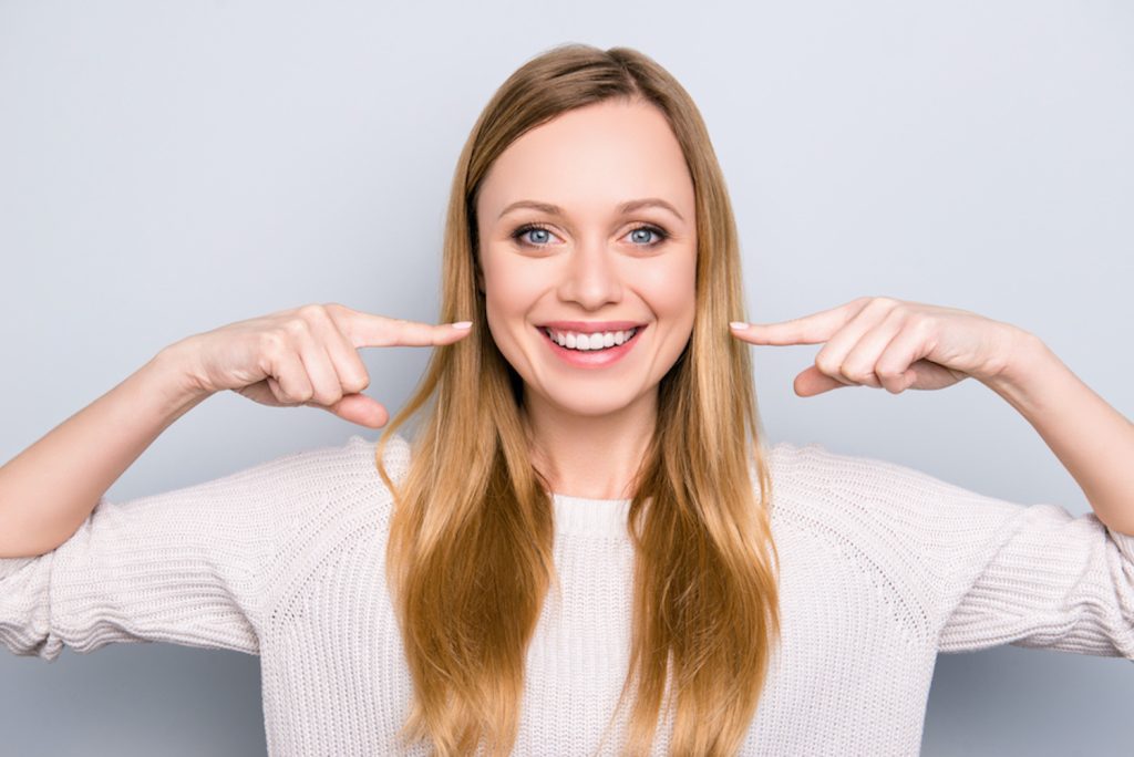 Blonde woman smiles and points to her teeth after finishing her orthodontic treatment